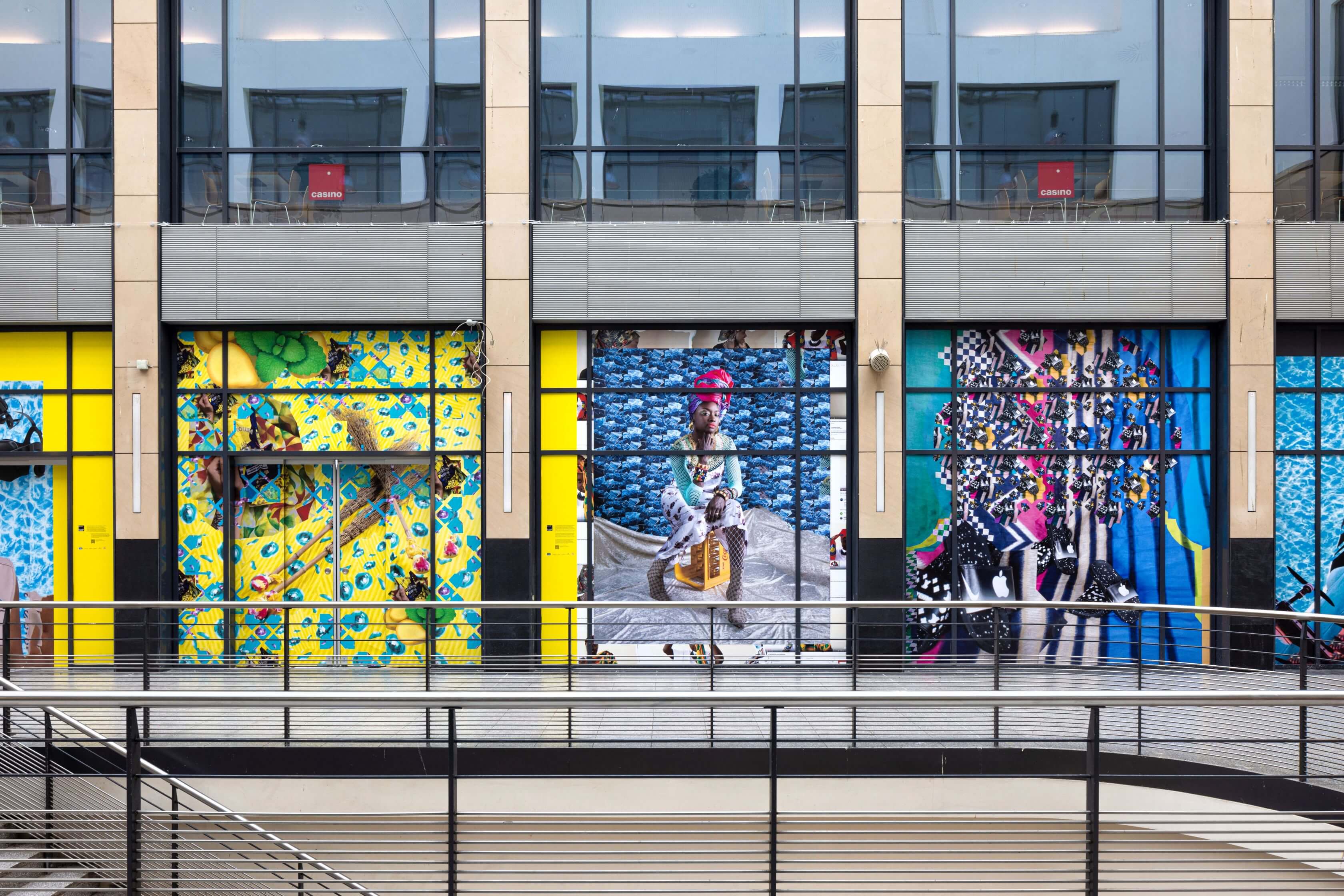 Die Fotografie zeigt einen Ausschnitt des Mannheimer Hauptbahnhofs mit drei digitalen Collagen aus der Serie Tools for Conviviality. Die Collage in der Mitte zeigt das Portrait von Awa, die in einer selbstbewussten Pose auf einer gelben Coca-Cola-Kiste sitzt. Links und rechts von ihr sind weitere bunte Collagen mit verschiedenen sich wiederholenden Mustern zu sehen