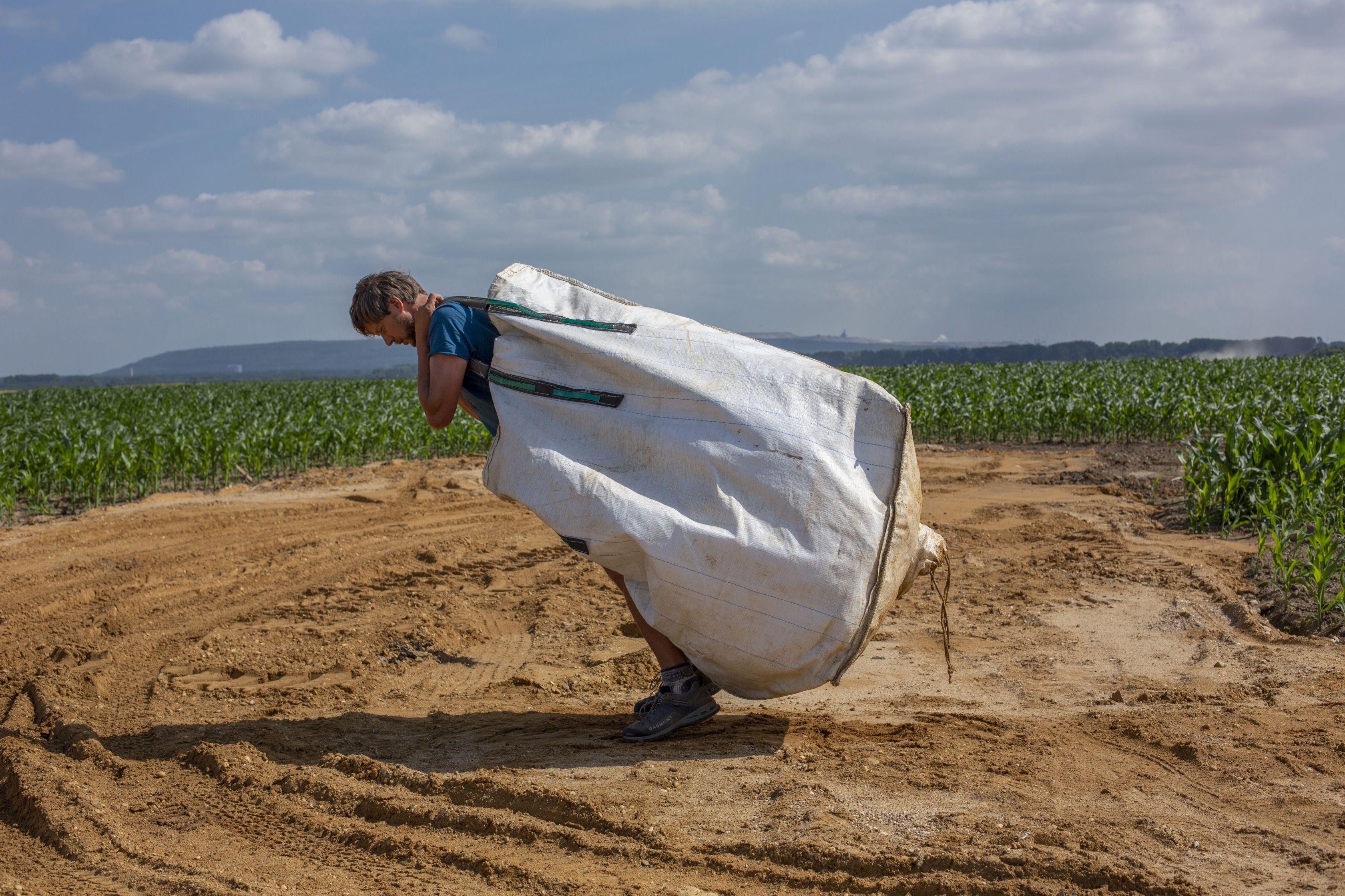a man stands bent over in a field carrying a white sack on his back